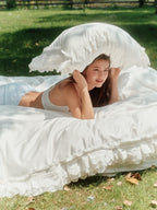 Woman holding a Crème ruffle pillowcase while lounging on the matching bedding set